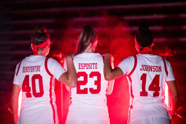 photo of (from left) Sophie Beneventine (CAS’24), Emily Esposito (COM’22), and Sydney Johnson (CAS’23) wearing their basketball jerseys, facing away from the camera, and standing in a dark room; a red light showcases their profiles. Sophie and Sydney stand with their hands resting on Emily's shoulders.