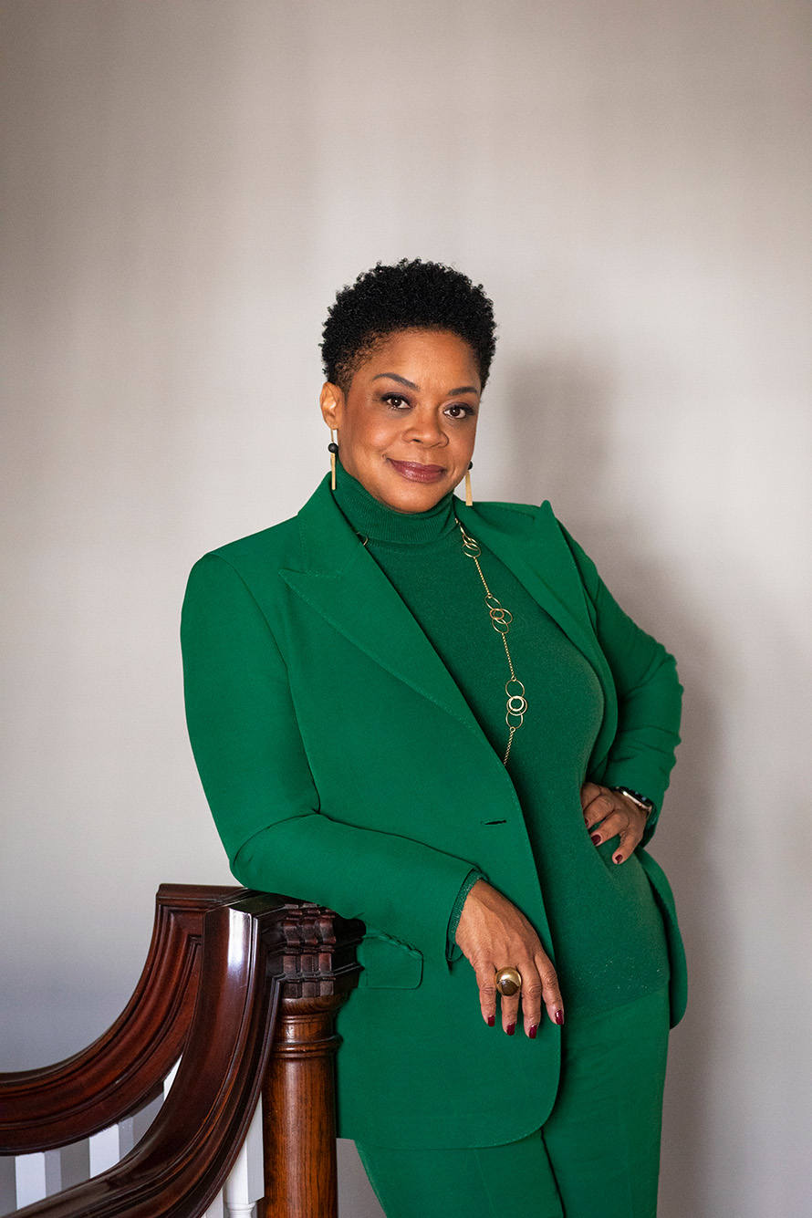 photo of Crystal Williams smiling and posing in front of a studio backdrop. She wears a dark green fitted suit and turtleneck an leans slightly against a wooden chair and her other hand rests on her hip