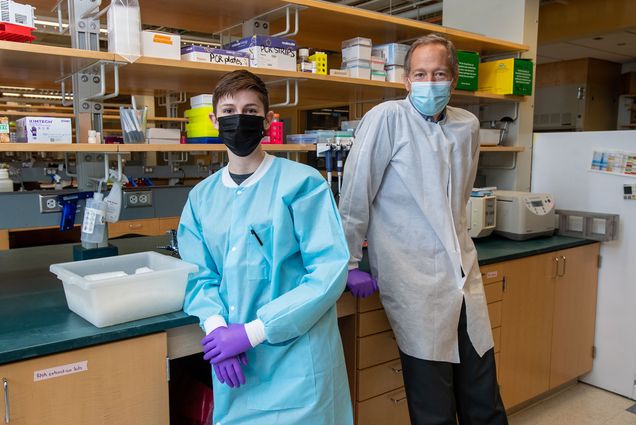 photo of Third year PhD student Jackie Turcinovic, left, and John Connor, PhD in the Connor Lab. Turcinovic wears a light blue PPE layer over her clothes and purple gloves while Connor wears a grey lab coat and leans back onto the counter.