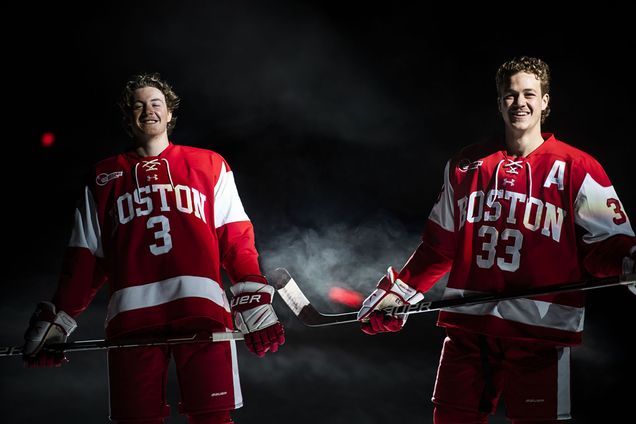 Photo of Ty and Tristan Amonte posing with hockey sticks in their red BU jerseys as a small white cloud of smoke rises behind him. The background is black and the brothers are dramatically lit.