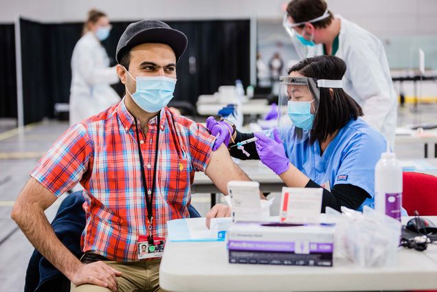 Photo of Mohsan Saeed, a School of Medicine assistant professor, getting a first vaccine dose in January of 2021. He wears a red and blue plaid shirt and gray hat, while the nurse, in a face mask and shield gives him a vaccine in his left arm.