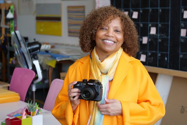 Photo of Linda Calhoun (COM’80), a middle-aged Black woman with light brown, curly, shoulder-length hair. She wears a long, yellow, draping blouse and holds a DSLR camera as she stands in what looks like a studio.