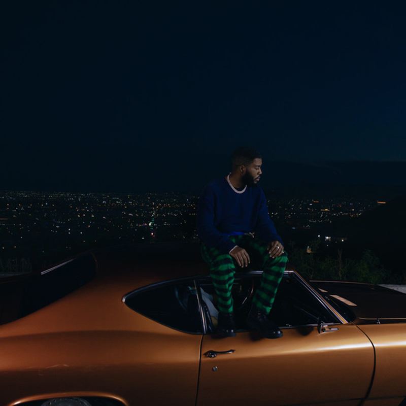 Album Cover of Khalid's "Scenic Drive". Image shows Khlad sitting in a burnt orange car as it overlooks a nighttime city view
