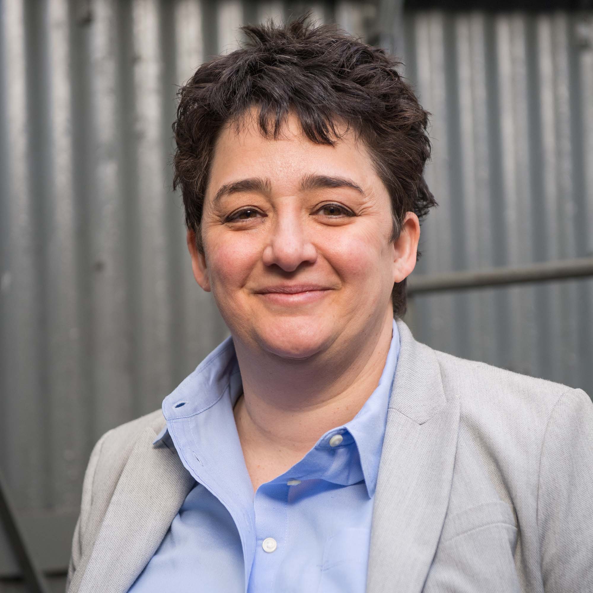 photo of smiling Merav Opher. Opher wears a light blue collared shirt and light gray blazer.