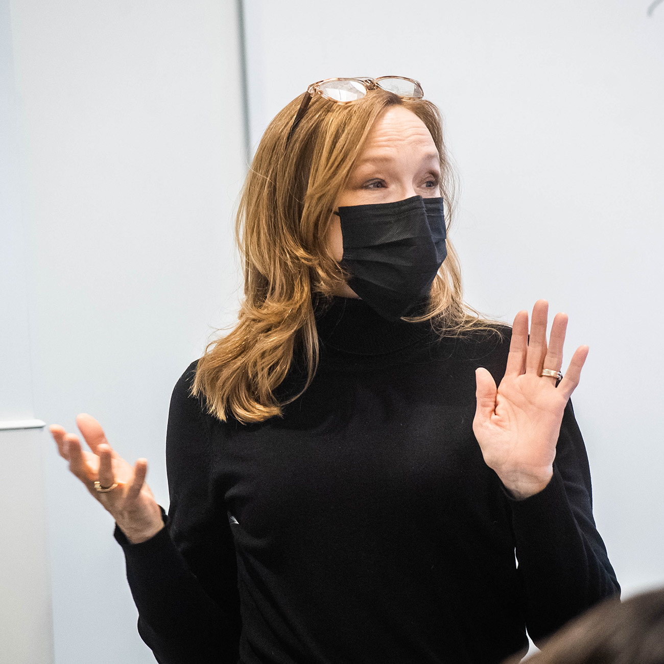 Photo of CAS Writing Program lecturer Jamie Robertson, who wears a black face mask and black turtle neck. She has blonde shoulder length hair and balances glasses on her head.