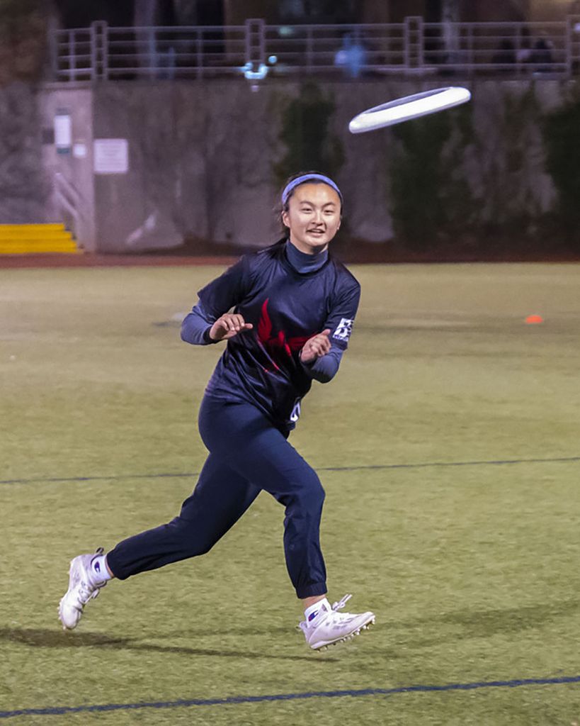 Photo of Liu during an ultimate game. She wears a dark blue outfit and headband and smiles slightly as she prepares to catch a white frisbee that is mid-flight.