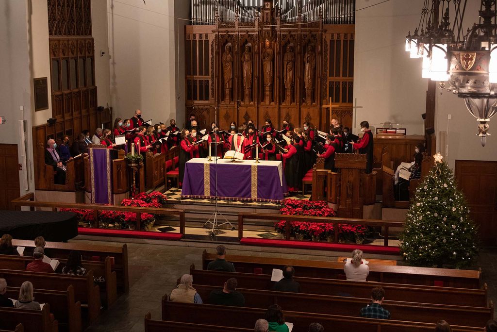 Photo of the 48th Service of Lessons & Carols on Friday, December 10, 2021 at Marsh Chapel. The chorus leader stands behind an altar covered with purple close and directs the rows of singers dressed in red robes and holding song books. In the foreground, the backs of peoples' heads in the audience are seen.