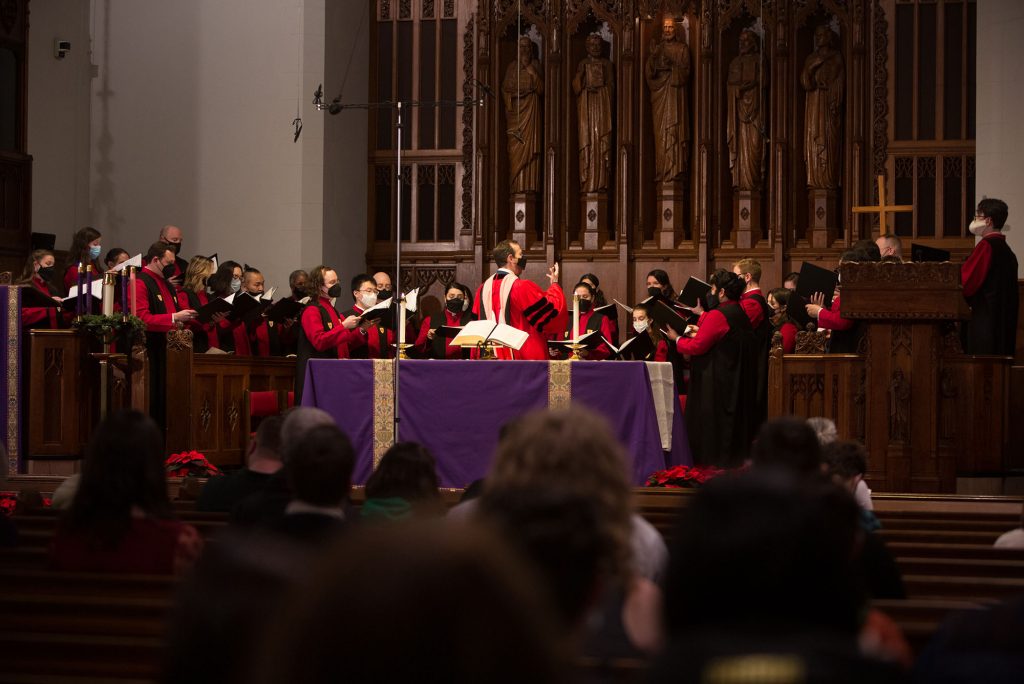 Photo of the 48th Service of Lessons & Carols on Friday, December 10, 2021 at Marsh Chapel. The chorus leader stands behind an altar covered with purple close and directs the rows of singers dressed in red robes and holding song books. In the foreground, the backs of peoples' heads in the audience are seen.