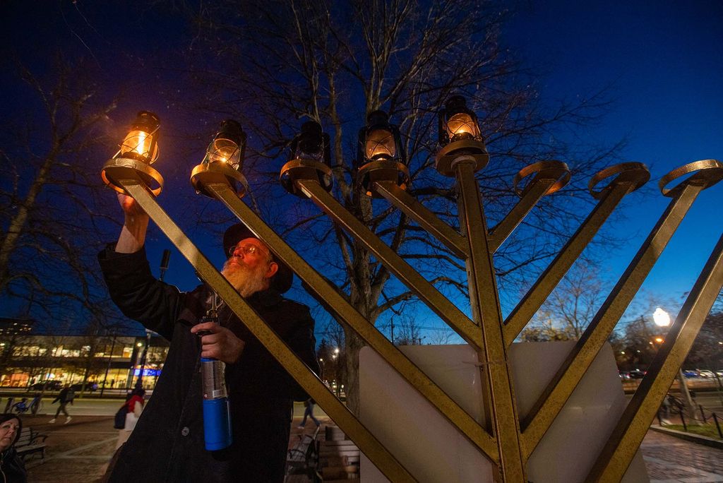 Photo of Rabbi Shmuel Posner lighting a large menorah outside the GSU Dec 1, the fourth night of Hanukkah. He has a long gray beard and a black hat, and dark blue sky is seen behind him.