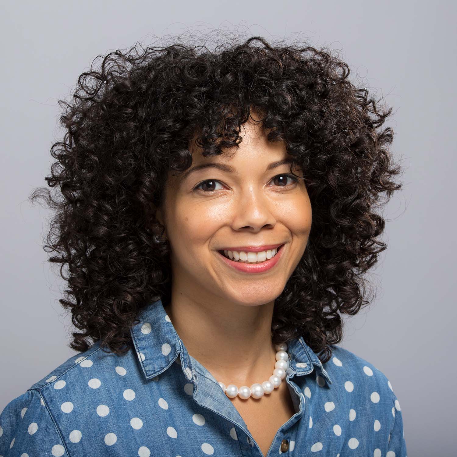 Photo of Amber Payne who wears a blue white-polka dot blouse. She smiles, has brown curly hair, and wears a pearl necklace.