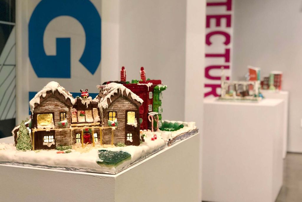 Photo of an elaborate ginger bread house with a frosting, snow covered roof on a white pedestal. The house has two floors and lights inside. 