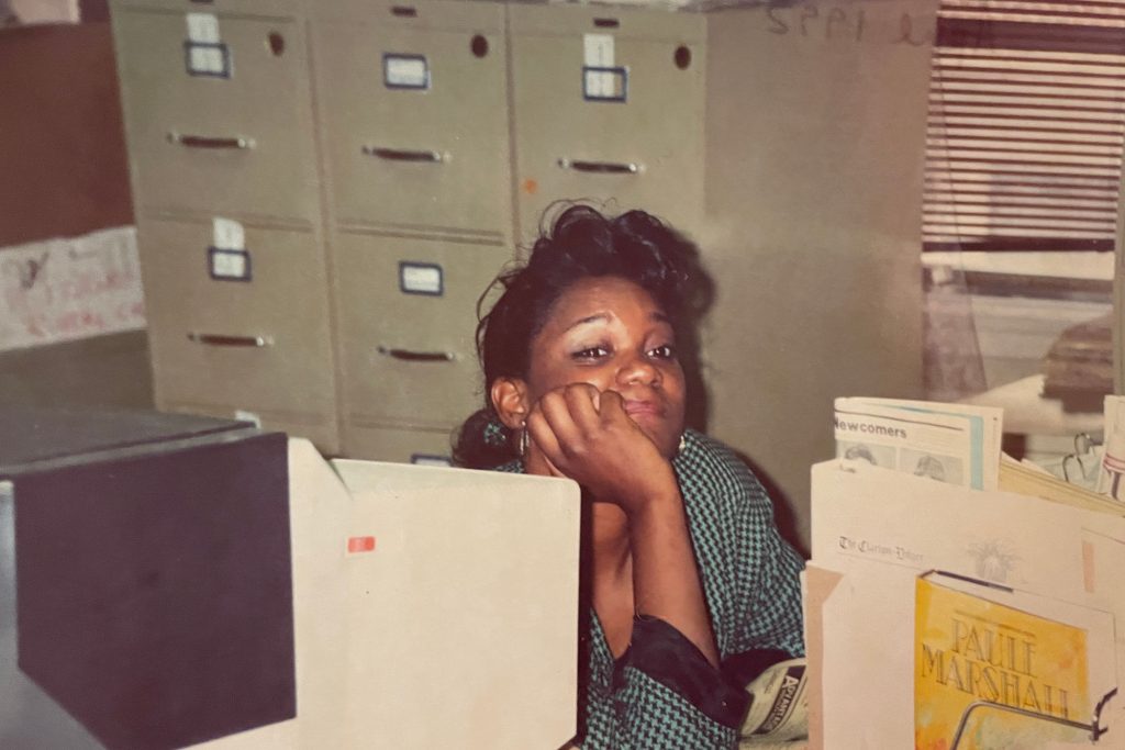 Photo of Douglas as a young journalist. She wears a teal pull over and rests her hand on her cheek. She sits behind a desk with file cabinets in the background. The photo appears to have been taken of a printed photo.