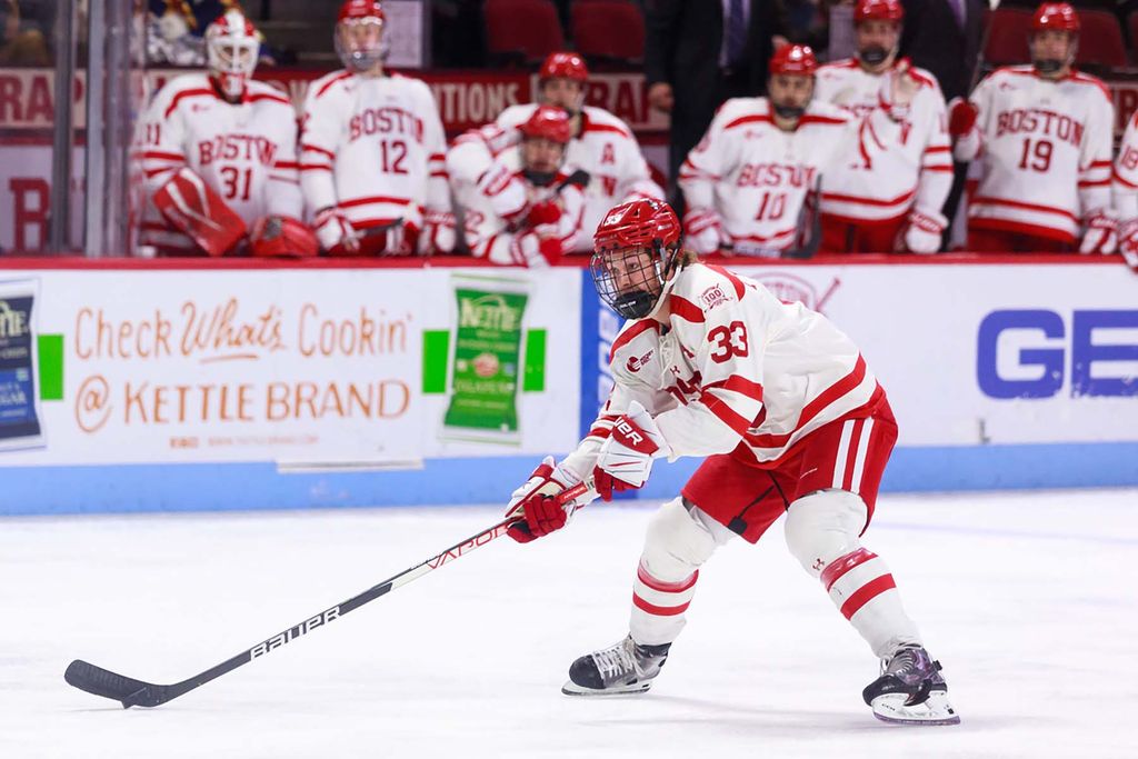 Photo of Ty Amonte (SHA’22) handling the puck during a game. He wears a white and red jersey and his teammates look on from the side line.