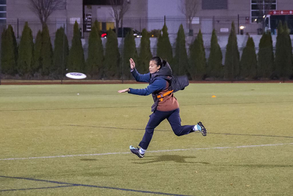 Photo of Elizabeth Wu wearing a jacket and running across a turf field to catch a white frisbee mid-flight.