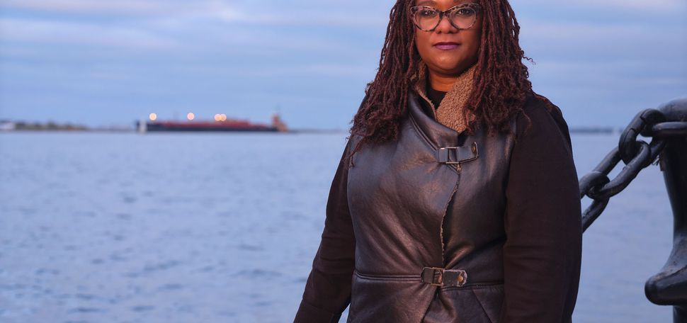 Photo of Deborah Douglas along boston harbor. She wears a black leather jacket, has shoulder-length, crimson locs, and rests her hand on a large middle chain fence. Behind, the harbor at twilight is seen.