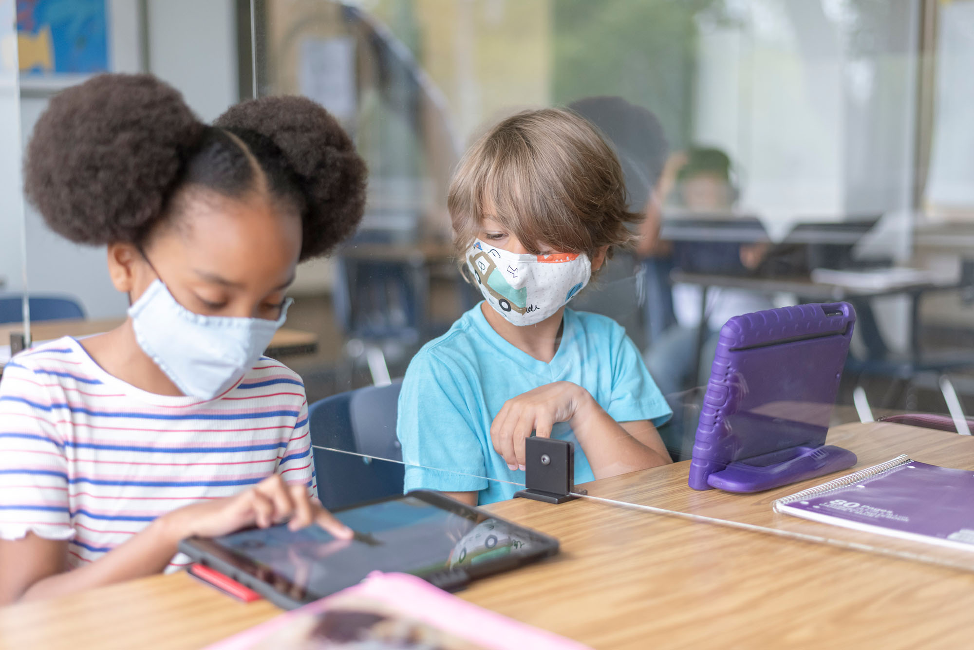 two school children sit next to each other with a plastic divider between them and look at a tablet together