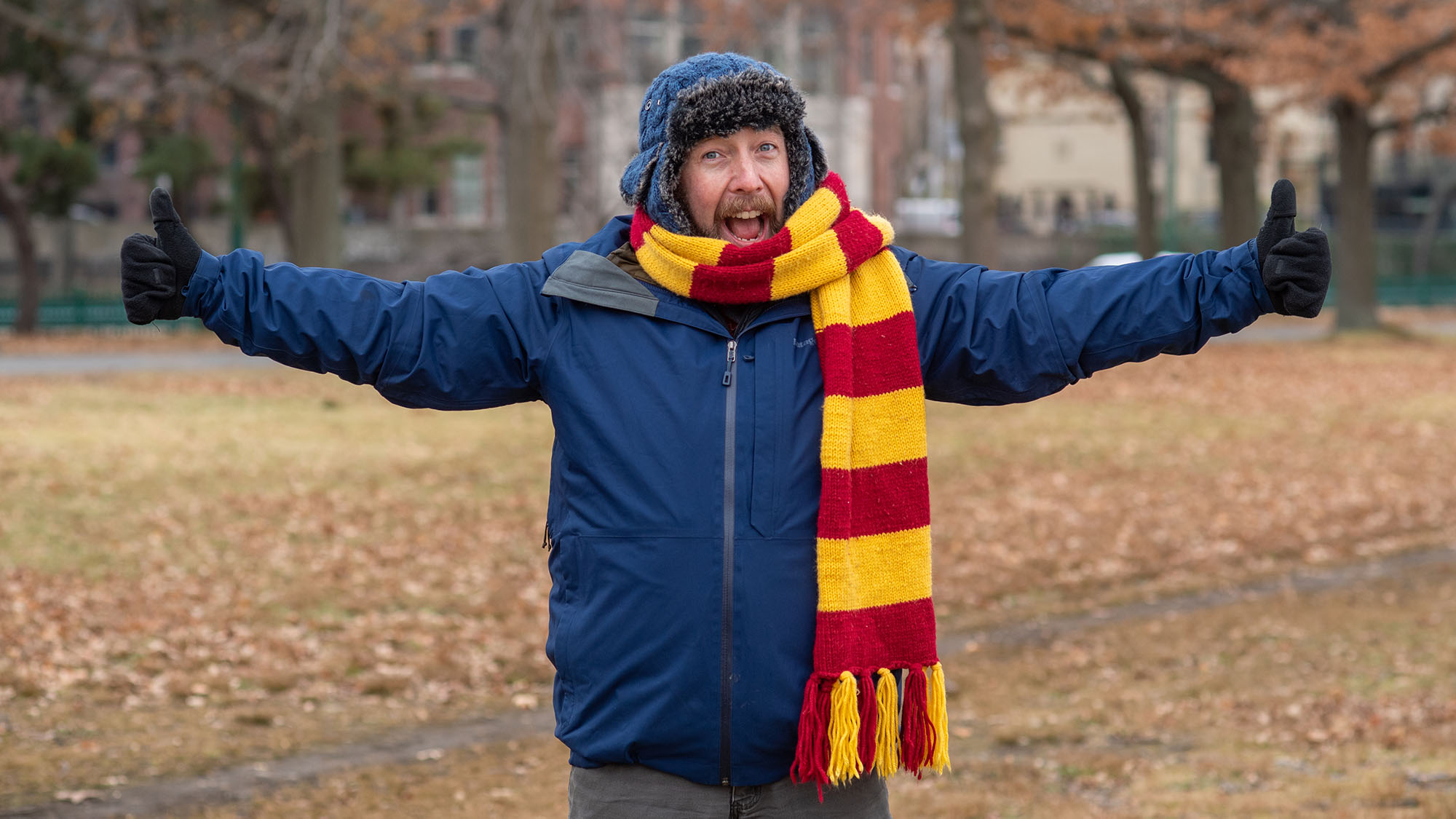 Photo of Hutch Hutchinson, a veteran New England outdoorsman, wearing a yellow and red scarf, blue hat with panels, and blue winter coat. He smiles, extends his arms joyfully, as he explains tips for how to dress for winter.