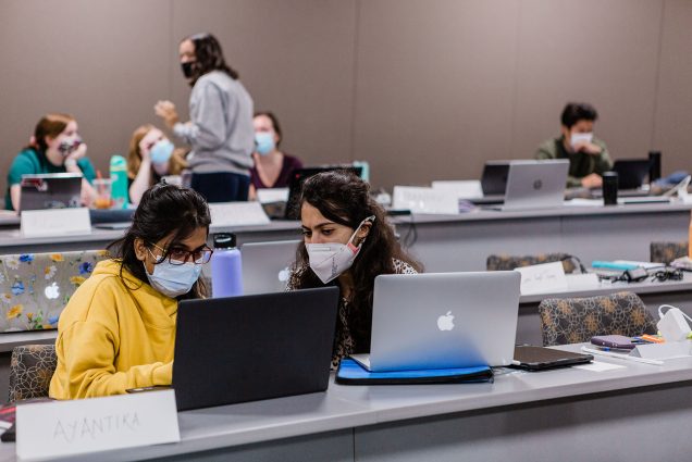 Photo of two students, sitting in a classroom with their laptops open. They wear face masks and one looks at the other's laptop. Students at the back of the classroom talk in a small group.
