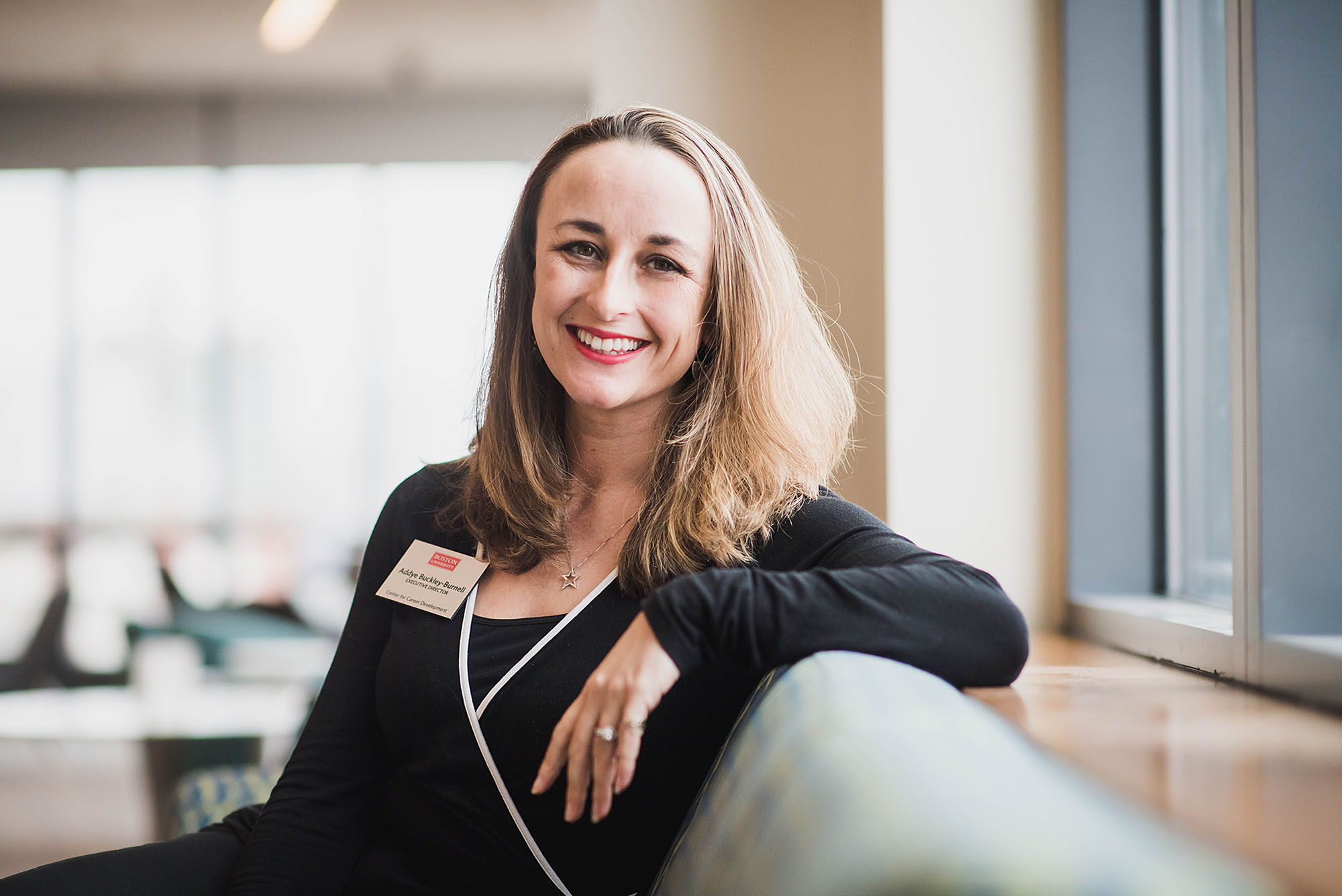 photo of Addye Buckley-Burnell posing for a photo. A white woman with dirty blonde hair sits and is turned to look at the camera as she smiles in a bright room