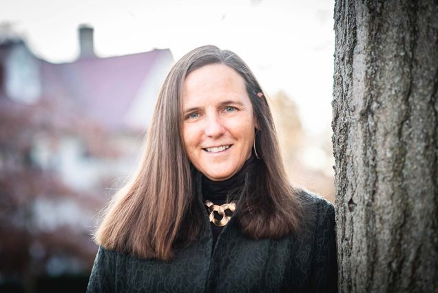 Photo of BU law professor, Katharine Silbaugh on January 4, 2022. She clasps her hands, smiles, and leans against the trunk of a tree. She has brown hair and wears a blue, speckled jacket over a black blouse.