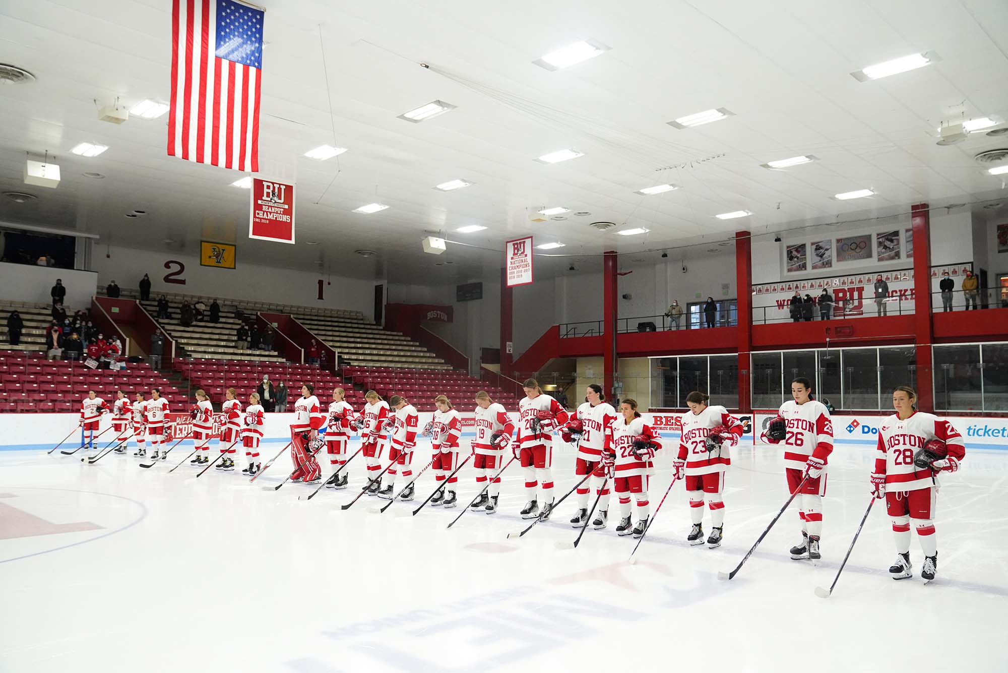Photo of the BU Women's hockey team standing in a row across the ice with their helmets off and white and red home jerseys on, during the national anthem before the Jan 15 game at Walter Brown Arena. The American flag hangs from the ceiling.