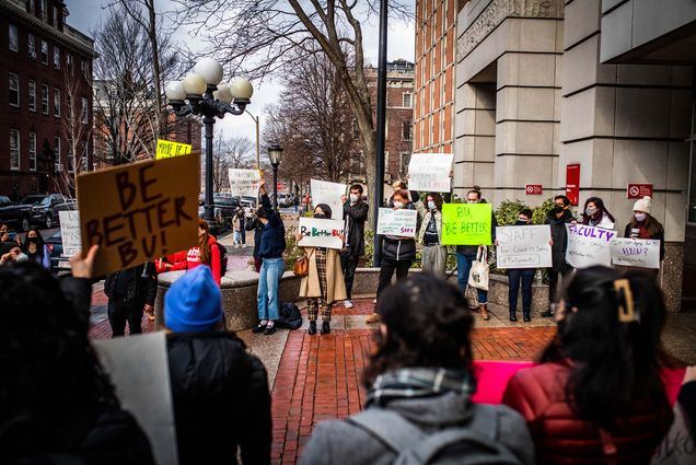 photo of School of Social Work students staging a walk-out outside of 1 Silber Way to protest BU's COVID-19 safety policies. They hold a variety of multicolored signs with the hashtag "#BeBetterBU".