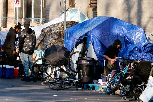 Photo of the tents of a homeless camp lining the sidewalk in area commonly known as Mass and Cass in Boston. A dark blue and light blue tent are placed together as people and bikes stand around them.