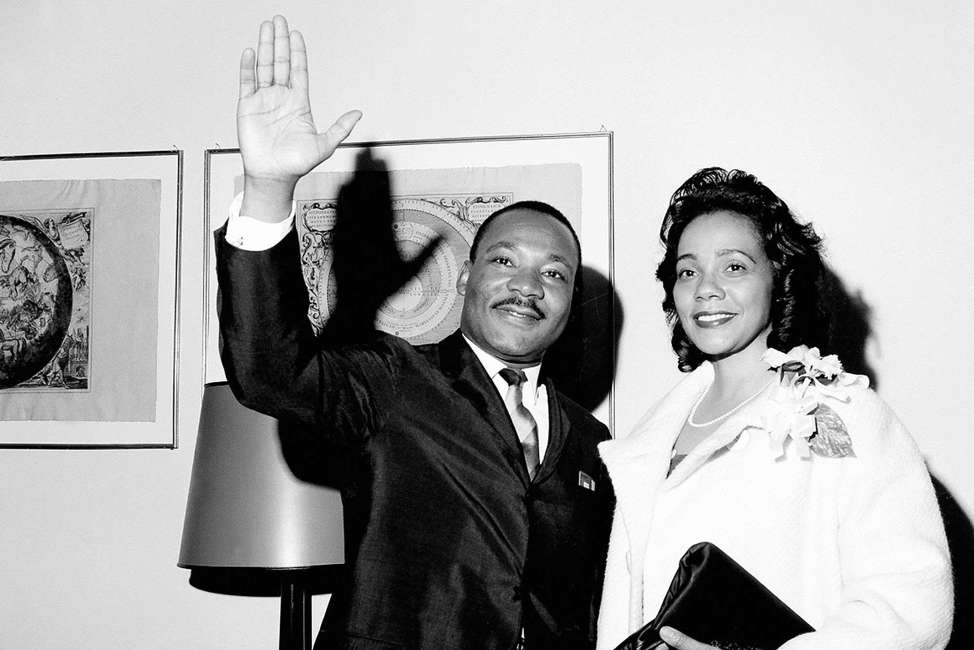 black and white photo of The Rev. Dr. Martin Luther King Jr. being accompanied by his wife, Coretta Scott King, as he appears at a press conference. His hand is raised and waving, as they both look on smiling