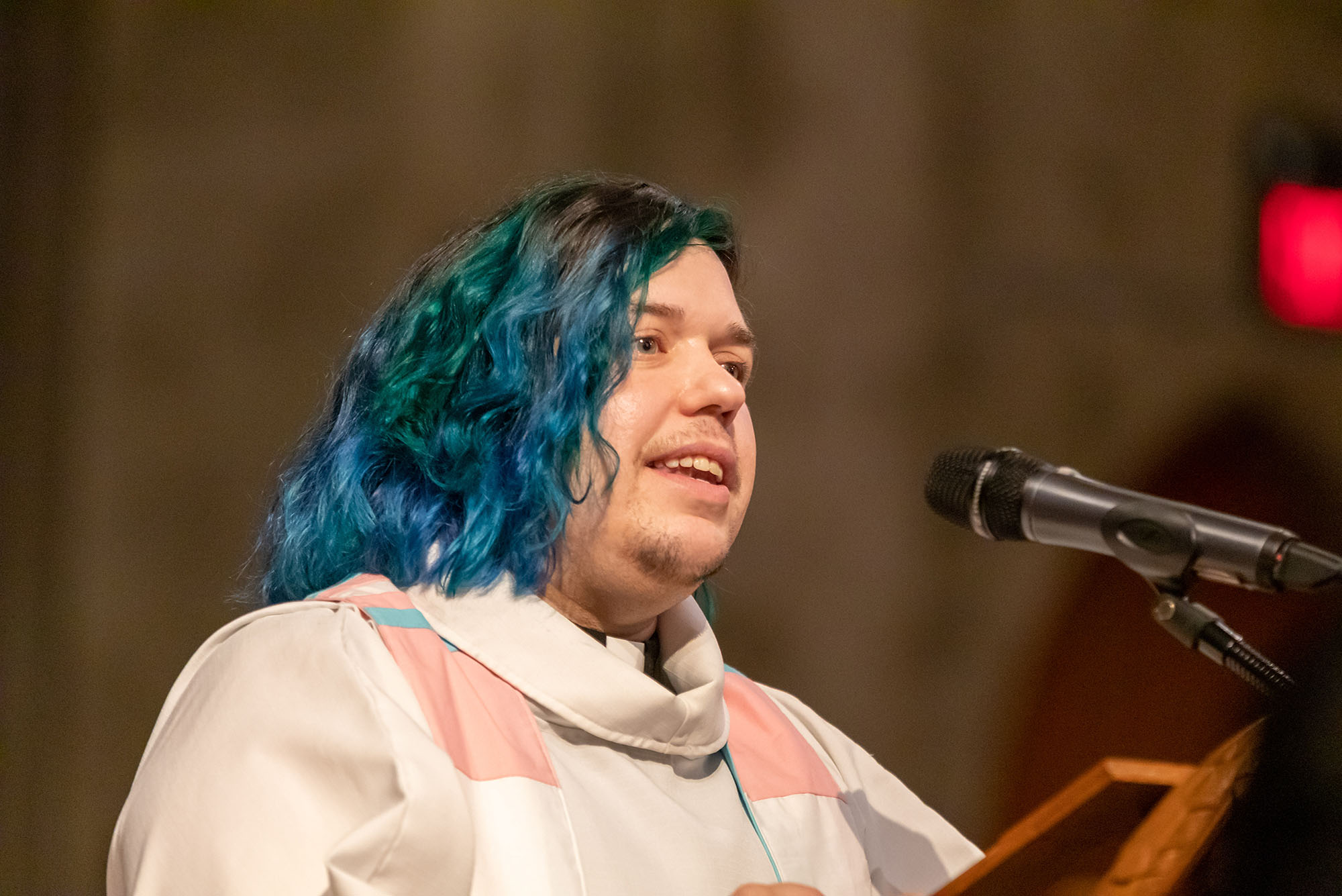 photo of Kori Pacyniak giving a sermon. They wear traditional priests robes and don blue hair.