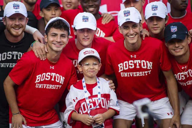 Photo of the BU men’s soccer team, wearing red shirts with 'Boston University' written in white, in a huddle around Team IMPACT member Pierce Sabin, a young child with a BU jersey and white BU hat, on October 16. All the young men and Sabin smile and celebrate their win.