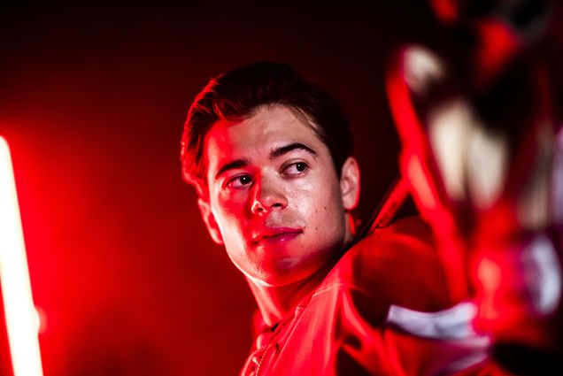 photo of BU men's hockey player Jay O’Brien, a white man, posing for a shot in a studio. He wears his hockey uniform and holds his stick behind his head in a dark studio with red, moody lighting.