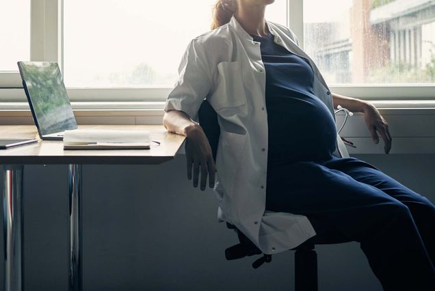 a pregnant doctor sits in an office chair at a desk with a stethoscope in their pocket