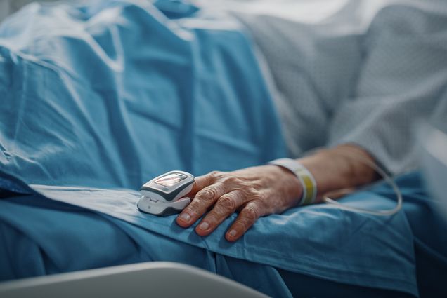 Photo of the hand of an elderly person resting in a hospital bed with a finger Heart Rate Monitor / Pulse oximeter on. The photo is zoomed in on the resting hand so the face of the patient is not visible.