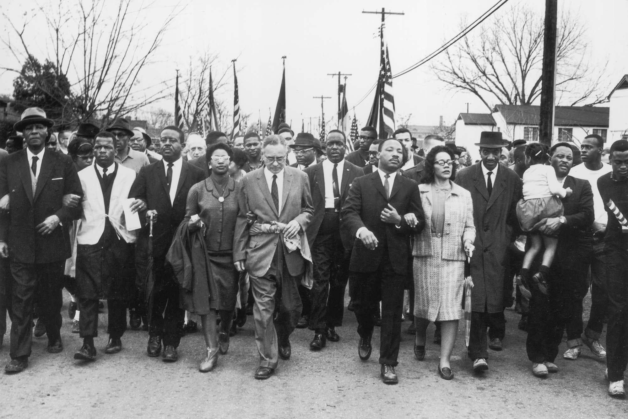 Black and white photo from March 1965, in which Martin Luther King, towards the right, in a suit, and his wife Coretta Scott King, in a hat and light pant suit, lead a black voting rights march from Selma, Alabama, to the state capital in Montgomery. Among those pictured are, front row, politician and civil rights activist John Lewis (1940 – 2020), Reverend Ralph Abernathy (1926 - 1990), Ruth Harris Bunche (1906 - 1988), Nobel Prize-winning political scientist and diplomat Ralph Bunche (1904 - 1971), activist Hosea Williams (1926 – 2000 right carrying child).