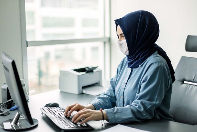 Photo of a young businesswoman in a dark blue hijab and blue button-down shirt working on a desktop computer while wearing a face mask.