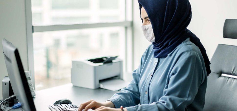 Photo of a young businesswoman in a dark blue hijab and blue button-down shirt working on a desktop computer while wearing a face mask.