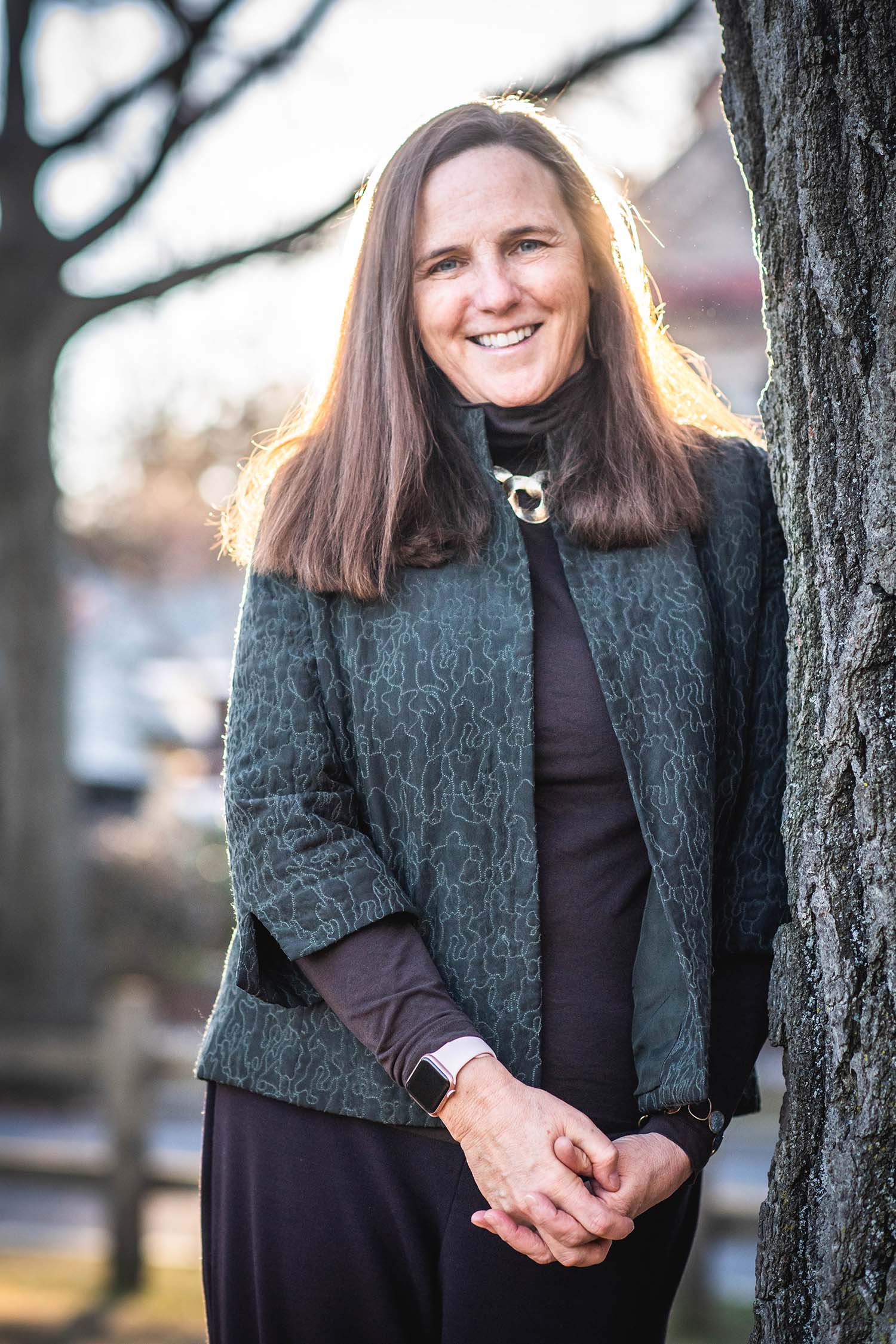 Photo of BU law professor, Katharine Silbaugh on January 4, 2022. She clasps her hands, smiles, and leans against the trunk of a tree. She has brown hair and wears a blue, speckled jacket over a black blouse.