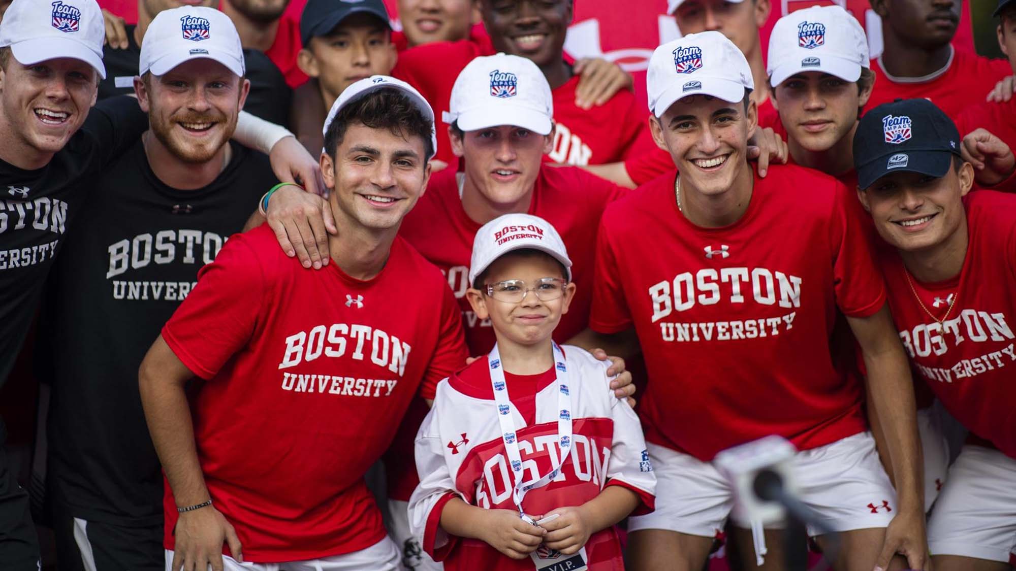 Photo of the BU men’s soccer team, wearing red shirts with 'Boston University' written in white, in a huddle around Team IMPACT member Pierce Sabin, a young child with a BU jersey and white BU hat, on October 16. All the young men and Sabin smile and celebrate their win.