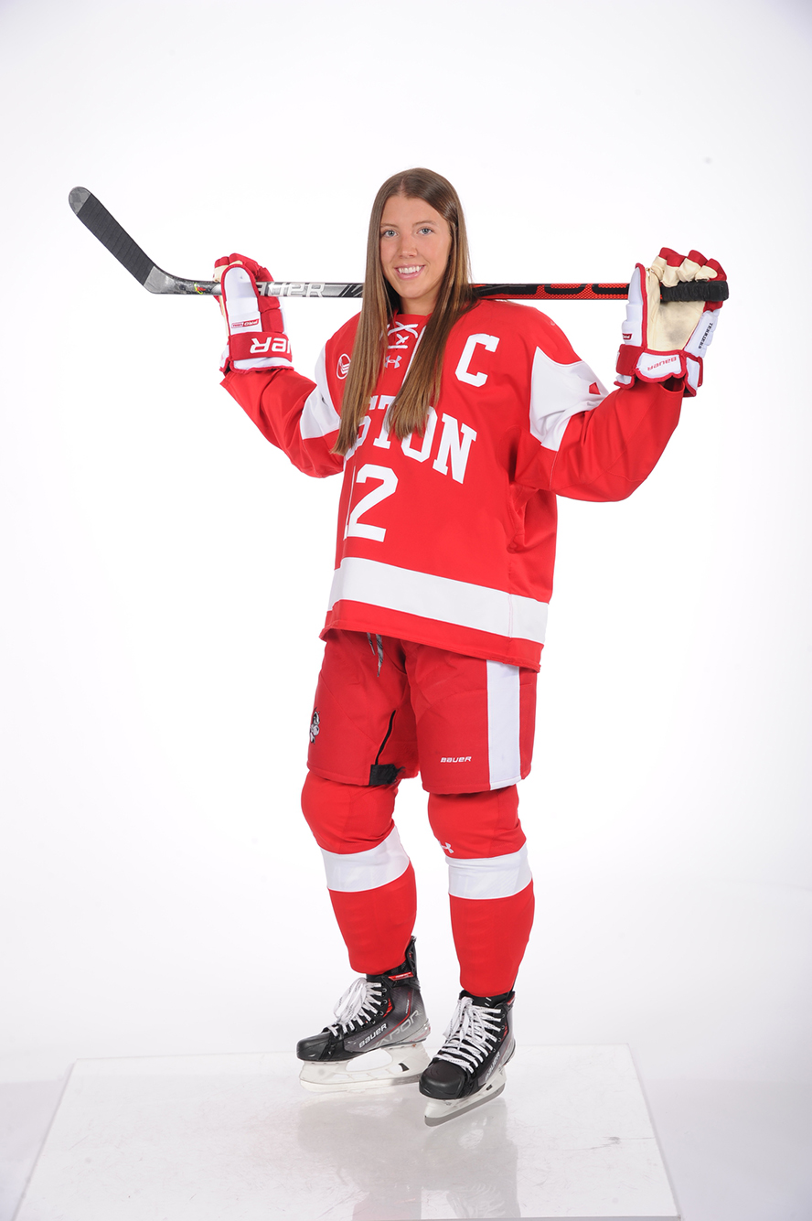 photo of Kaleigh Donnelly posing in her Boston University womens hockey uniform. She wears her skates and holds her hockey stick behind her head