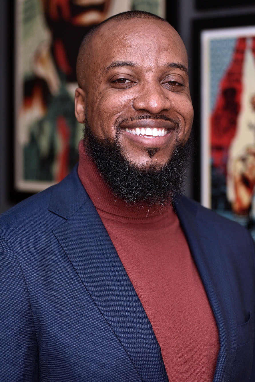 Headshot of Carrington Moore. He faces the camera and smiles, wearing a navy blazer and muted red turtleneck. 