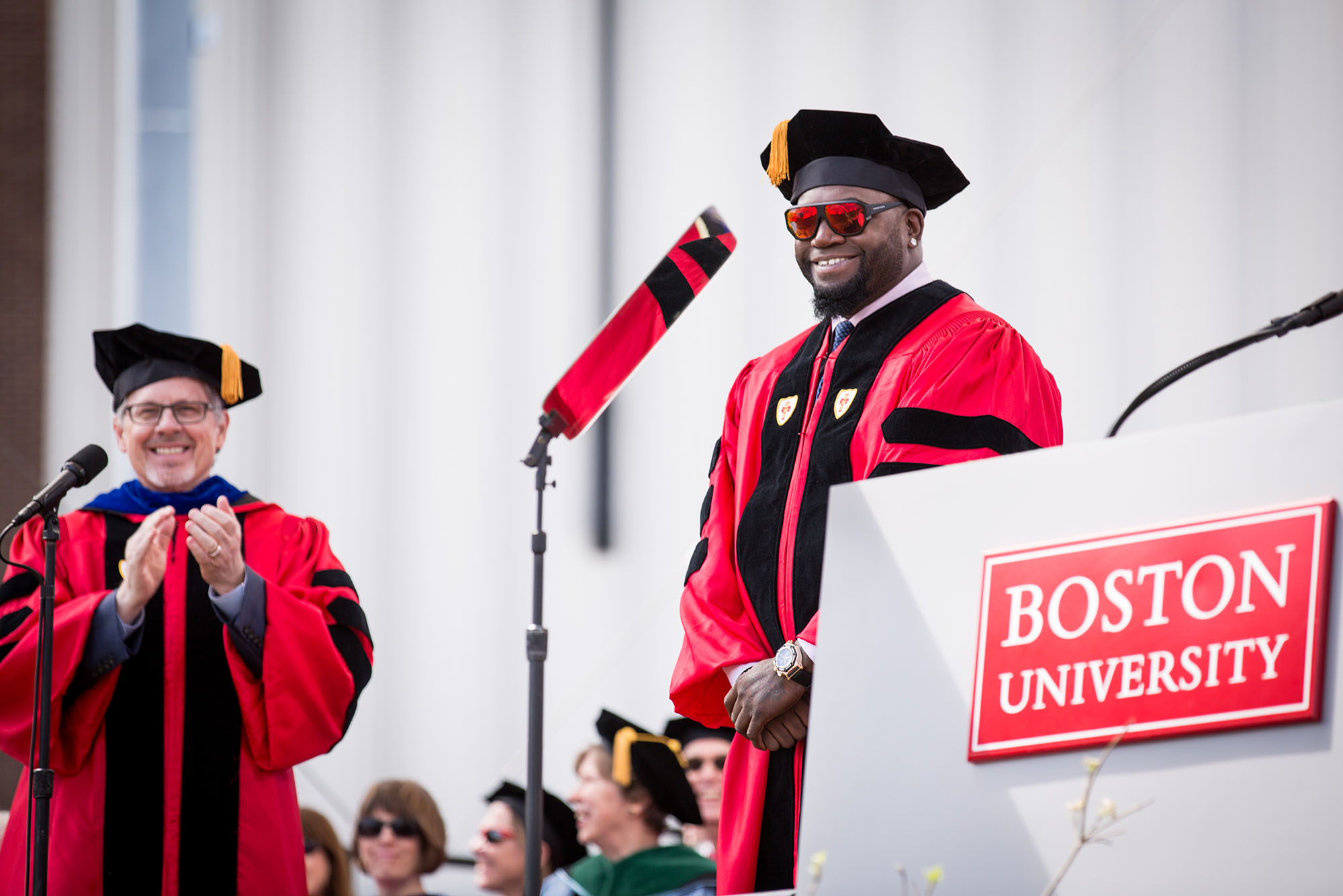 Photo of David Ortiz receiving an honorary degree during the 2017 Boston University Commencement on Nickerson Field on May 21, 2017. He wears a red graduation robe, black hat, and large sunglasses with red lens. He smiles wide with his hands clasped as a BU administrator claps in the background. In the right foreground, a podium with the red "Boston University" sign is seen.