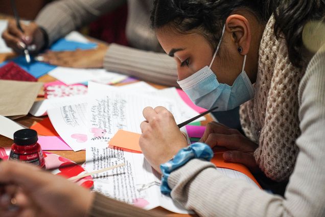 Photo of Simeon Seth (CGS'25) practicing basic words on a colored paper during the the January Calligraphy and Cocoa events at George Sherman Union sponsored by Student Activities Office. She wears a blue face mask and looks intently at the paper as she writes. Others writing can be seen behind her and the table is covered with colored paper.