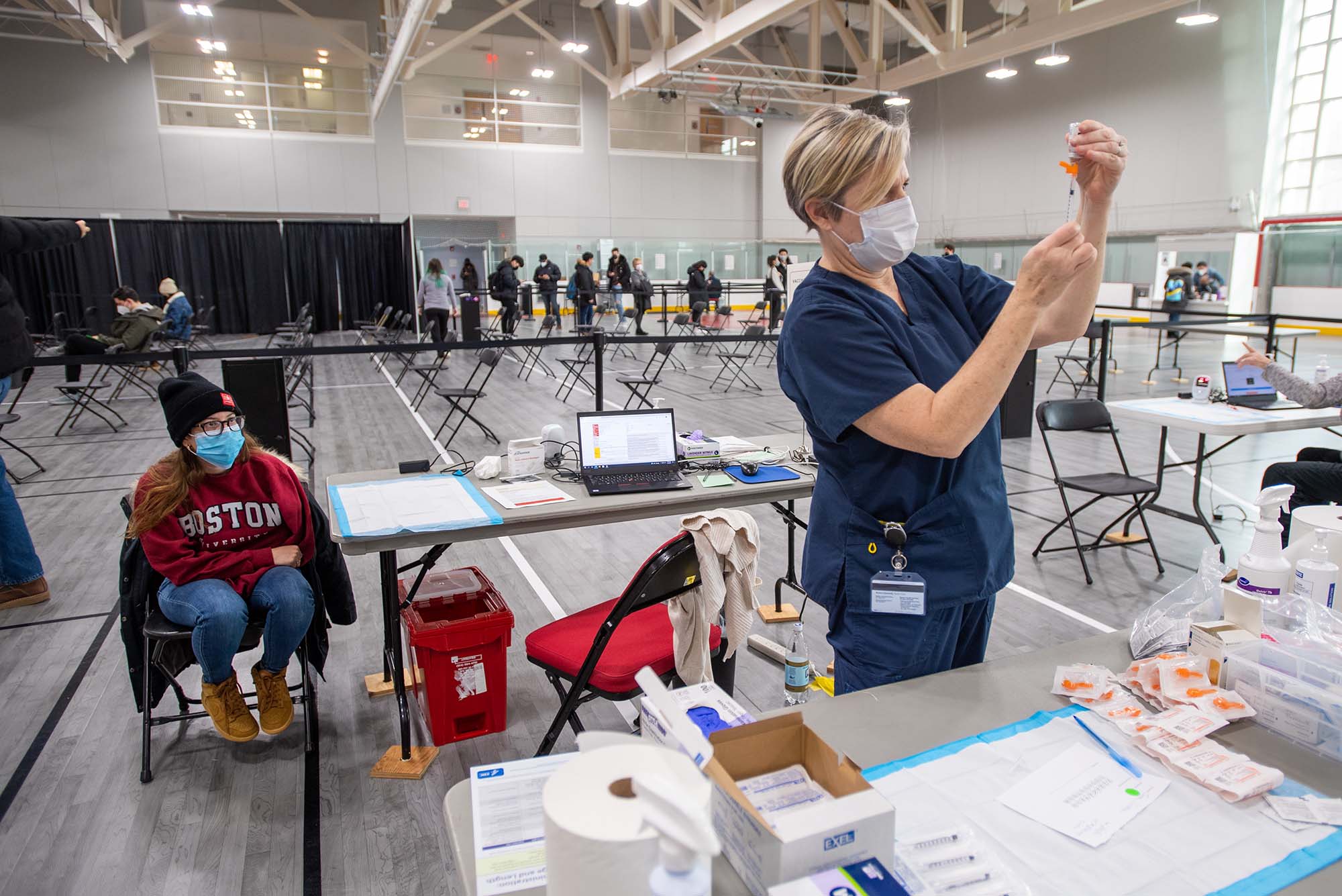 Photo of Healthway nurse Jane Twombly at right, preparing a dose for Jazmin Cruz-Flores (Wheelock’23) at left, at the Covid-19 vaccine booster shot clinic at FitRec January 25. Twombly wears dark blue scrubs and a face mask as she extracts the vaccine from the bottle. Cruz-Flores wears a red BU sweatshirt,hat, and a facemask.