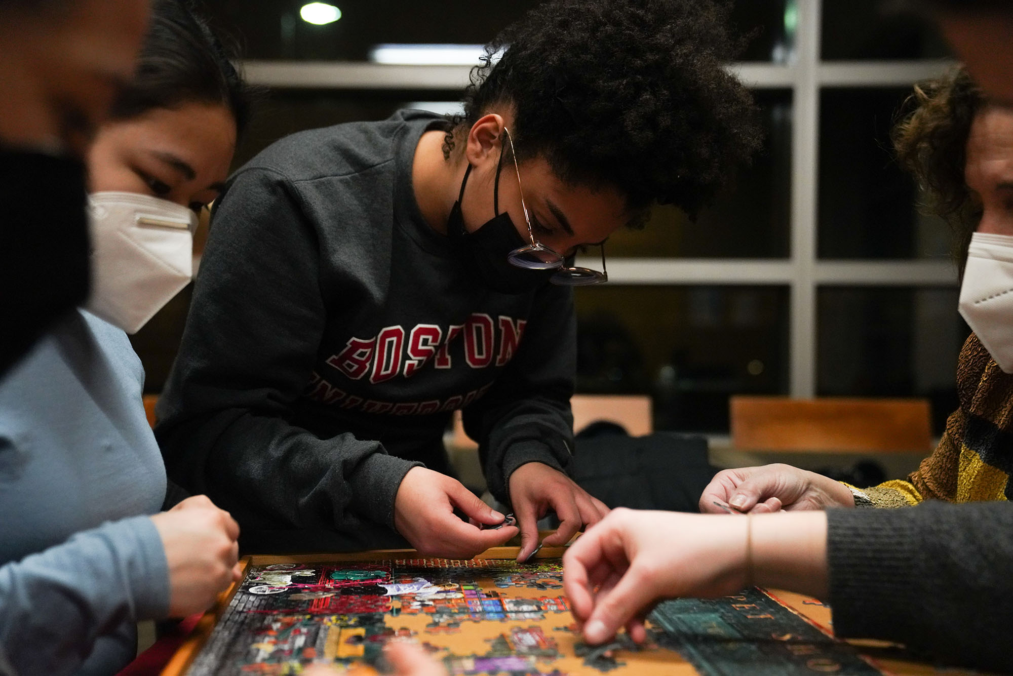 Photo of BU student Eliza Go Araujo (COM 24') putting pieces on the board while other students work on other parts of the jigsaw puzzle at the StuVi2 lobby in Jan 25. She wears glasses, a black face mask, and dark gray BU sweatshirt.