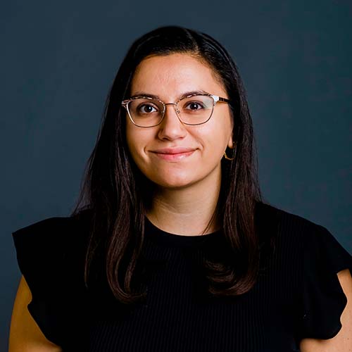 Photo of Mara Sasson, a young light-skinned woman with turtle glasses. She smiles and wears a black shirt with ruffly sleeves.