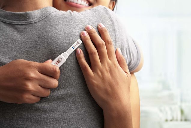 Photo of two partners, one dressed in a gray t-shirt, embracing as one holds a positive pregnancy test.