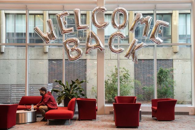 Photo of Lucas Amodio sitting on a red couch in the lobby of the LAW building. Behind him, large silver balloons in the shape of letters that spell "Welcome Back" hang behind him. The window behind him looks on to another building with a brick facade.