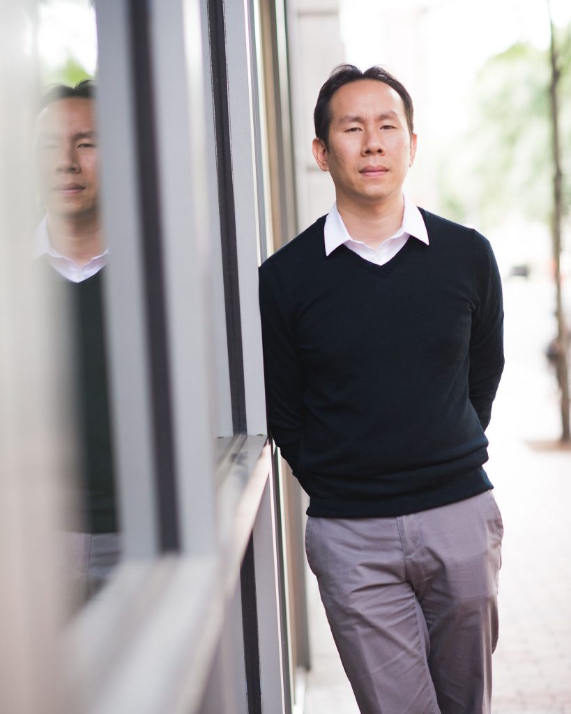 Portrait of Jerry Chen, whose recent study illuminates the sense of touch, wearing a black sweater and gray pants, and slightly smiling. He leans against a windowed wall.