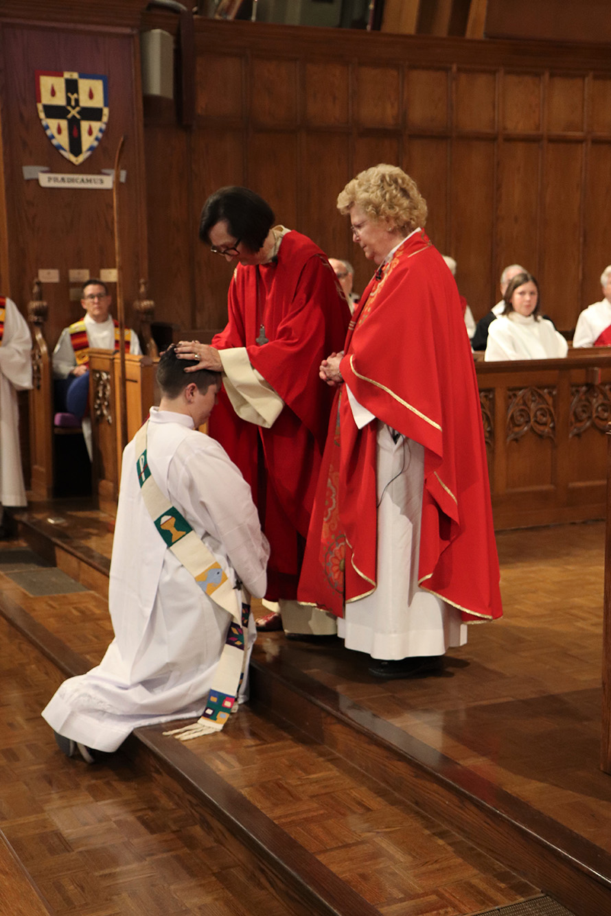 Photo of Kori Pacyniak being ordained. They kneel in front of two female priests in red robes. One rests her hands on Kori's head.