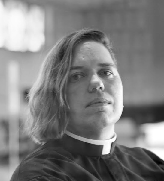 photo of Kori Pacyniak looking sternly at the camera as they wear the traditional black Catholic priest garbs.